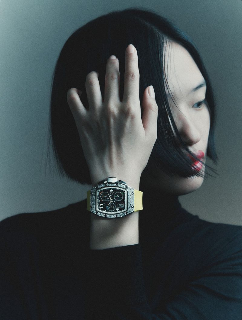 Close-up portrait of a model wearing a luxury wristwatch with a yellow strap and dark turtleneck against a plain background