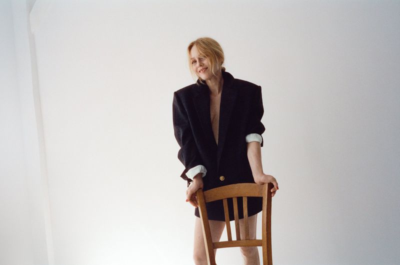 Vanessa Paradis wearing an oversized black blazer leaning on a wooden chair against a plain white wall