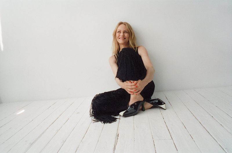 Vanessa Paradis seated on white painted wooden floor wearing black fringe trousers and black and white heels against a white wall.