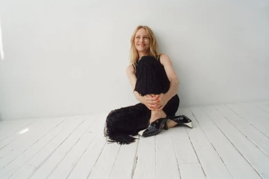 Vanessa Paradis seated on white painted wooden floor wearing black fringe trousers and black and white heels against a white wall.