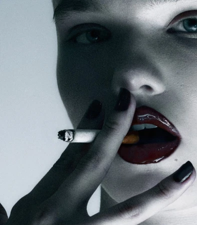Close-up portrait of a model with dark glossy red lips and dark nails holding a lit cigarette against a neutral grey background