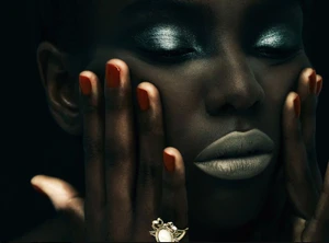 Close-up portrait of a Black woman with shimmering metallic green eyeshadow, neutral lips, orange nails, and a gold ring against a dark background.