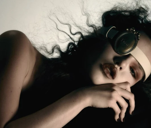Close-up portrait of a woman resting her face on her hand, wearing a headband and statement eyewear piece.