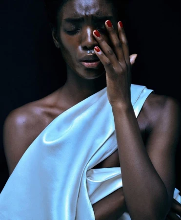 Black woman wearing a white draped garment with red fingernails touching her face against a dark background