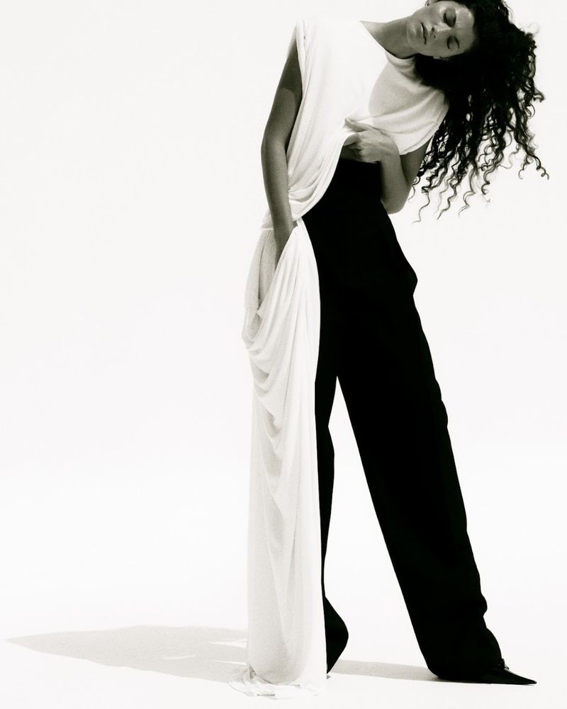 Model with dark curly hair wearing a white top and black trousers against a plain white background