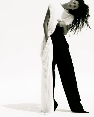 Model with dark curly hair wearing a white top and black trousers against a plain white background