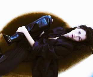 Model reclining in a round textured armchair wearing dark clothing and tall black boots.
