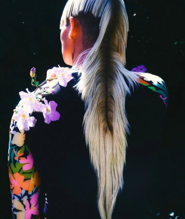 Model with long blonde ponytail and undercut hair viewed from behind wearing floral print sleeves and pale flowers resting on shoulder.