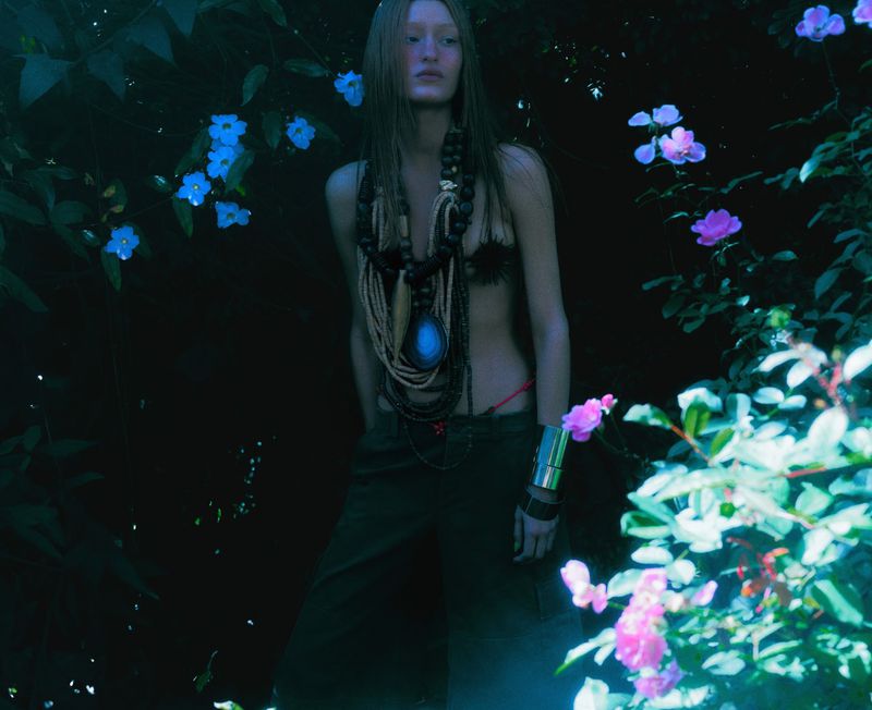 Model standing in a dark garden wearing layered statement necklaces, dark trousers, and a wide silver cuff bracelet amidst flowers.