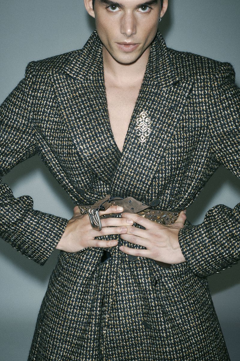 Portrait of a man wearing a patterned blazer with a studded belt against a grey background for Numero Netherlands.