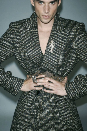 Portrait of a man wearing a patterned blazer with a studded belt against a grey background for Numero Netherlands.