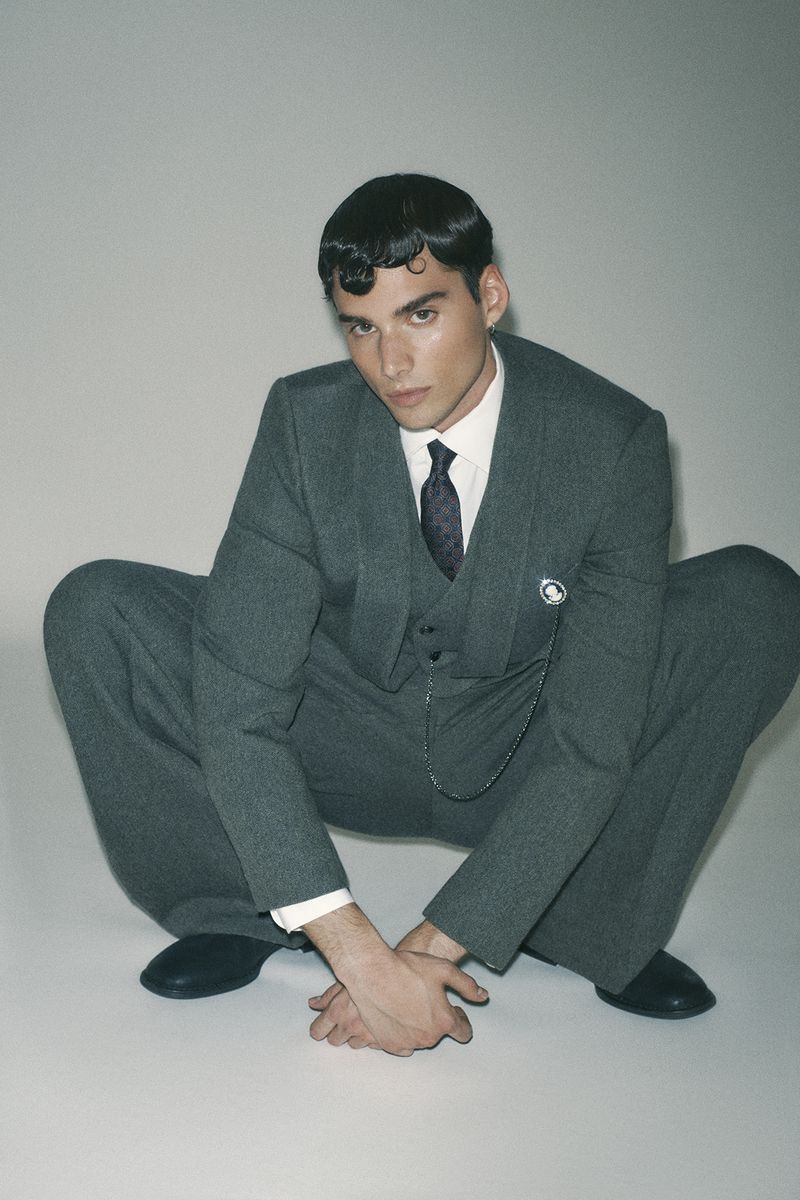 A man wearing a grey suit and tie crouches against a plain grey studio backdrop.