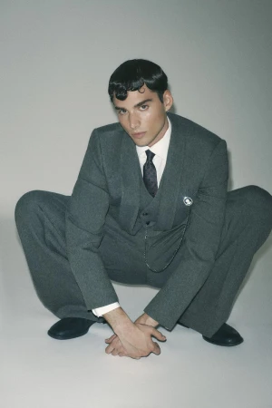 A man wearing a grey suit and tie crouches against a plain grey studio backdrop.