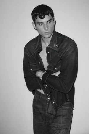 Monochrome portrait of a male model in a dark denim shirt and jeans against a plain background.