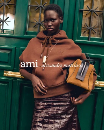 Model Chudier Billy wearing a brown hoodie and holding a handbag in front of a green wooden door for Ami F/W 26 editorial