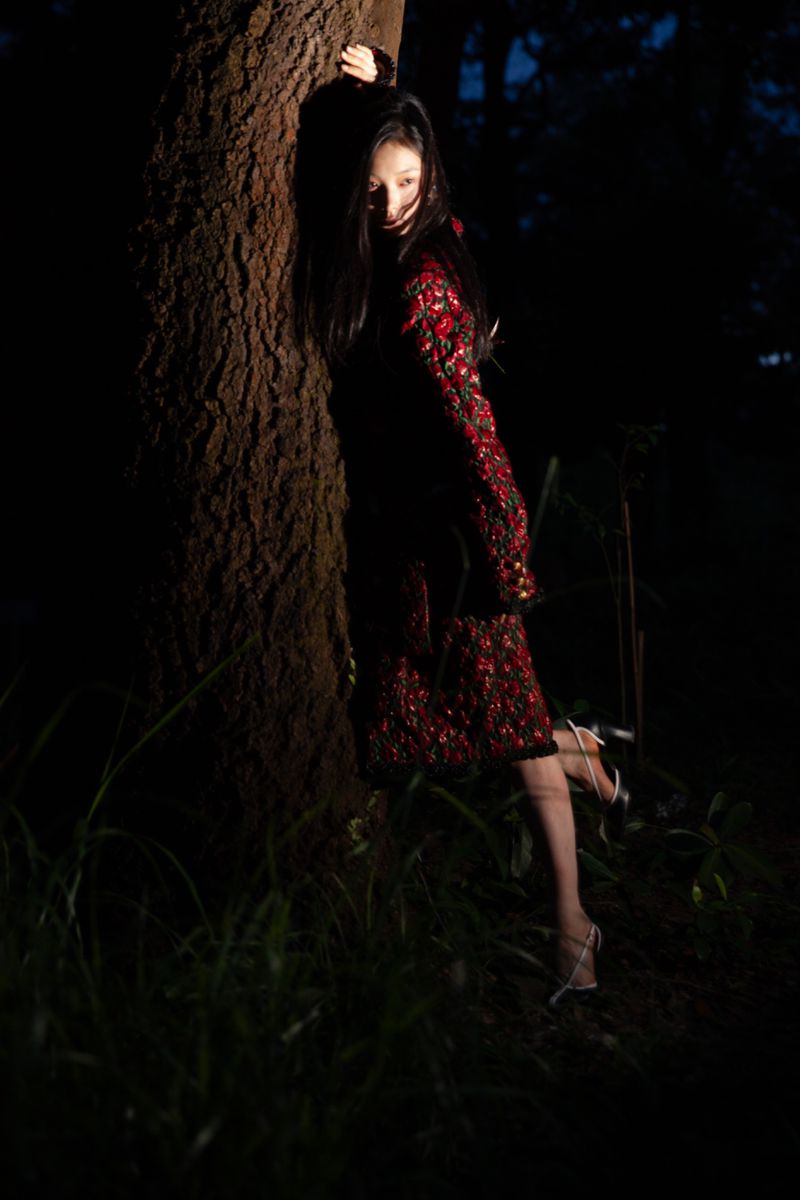 Model leaning against a large tree trunk in a dark forest wearing a red patterned dress