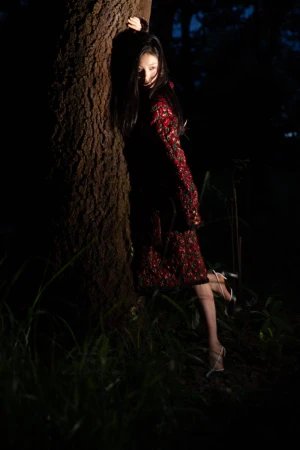 Model leaning against a large tree trunk in a dark forest wearing a red patterned dress