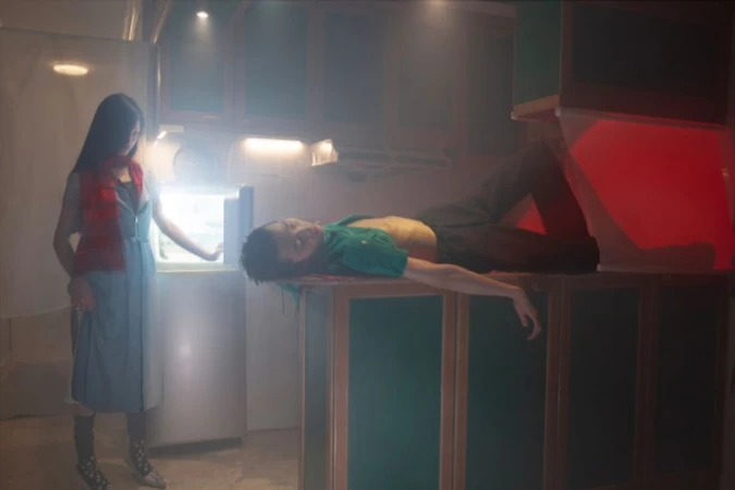 Two models in a dimly lit kitchen interior with hazy atmosphere, one woman standing by a lit refrigerator and a man lying on a kitchen counter.