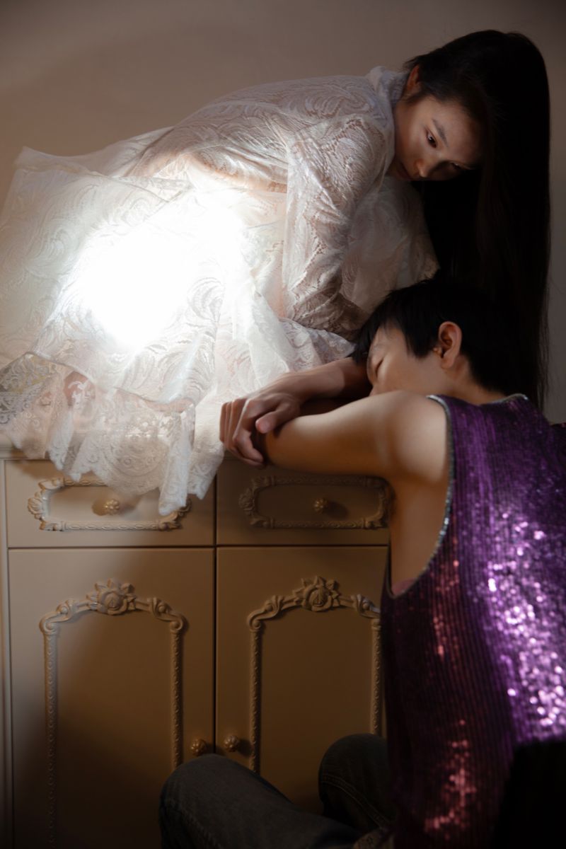 Two models posed together indoors with a woman in a white lace dress and a person in a sparkly purple top next to a cabinet.