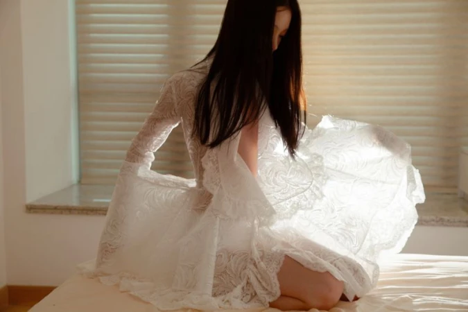 Model in white lace dress kneeling on a bed in a light-filled room for DNA Magazine Mexico