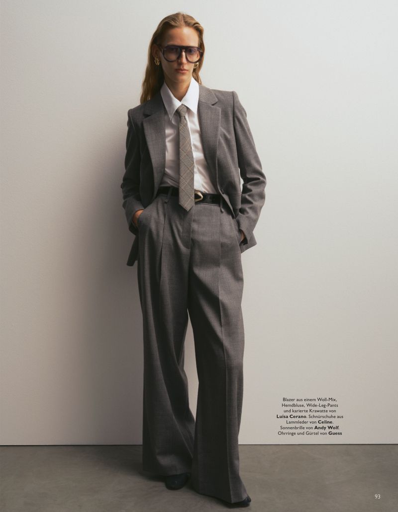 Model wearing an oversized grey pantsuit, white shirt, patterned tie and dark sunglasses against a plain studio backdrop
