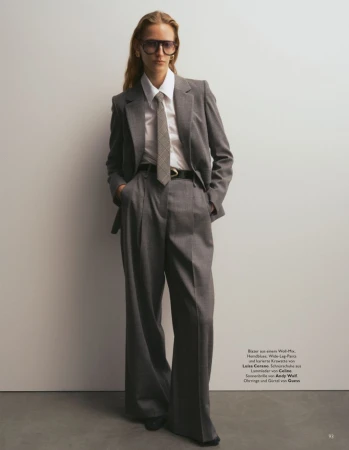 Model wearing an oversized grey pantsuit, white shirt, patterned tie and dark sunglasses against a plain studio backdrop