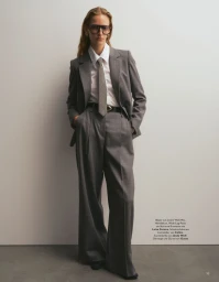 Model wearing an oversized grey pantsuit, white shirt, patterned tie and dark sunglasses against a plain studio backdrop