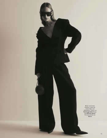 Model in dark oversized blazer and wide-leg trousers wearing large sunglasses against a plain studio backdrop