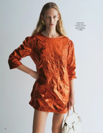 Model wearing a shiny orange metallic dress and holding a white handbag against a plain background