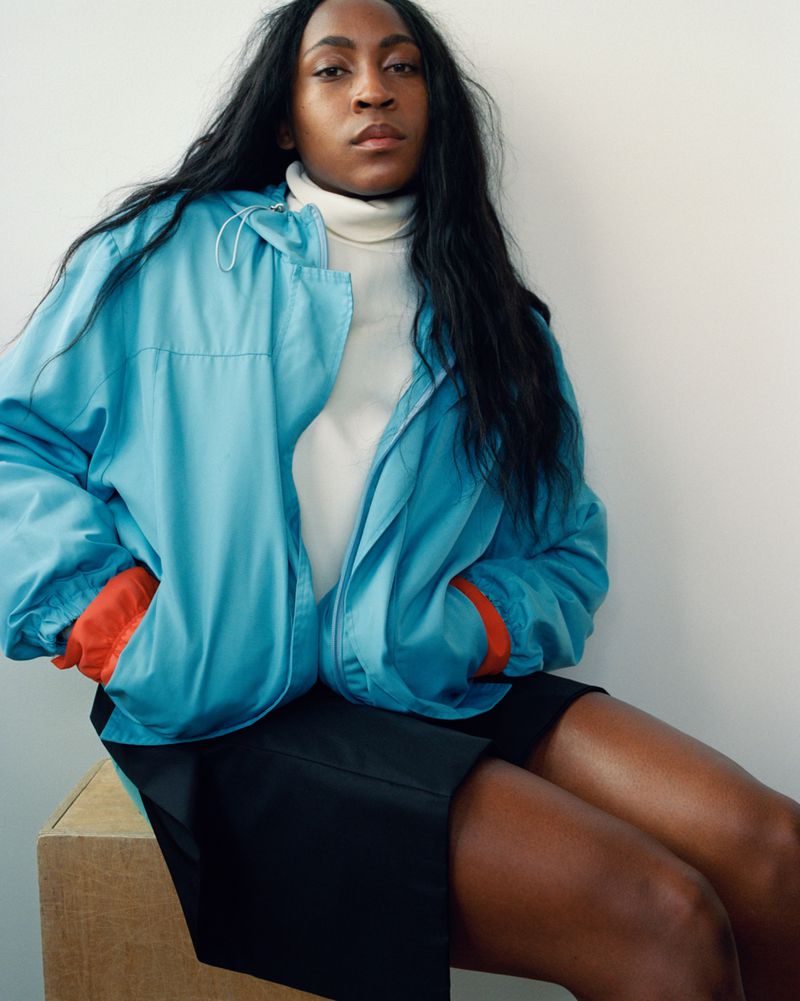 Coco Gauff seated in a blue zip-up jacket with orange cuffs, white turtleneck, and black skirt against a white wall.
