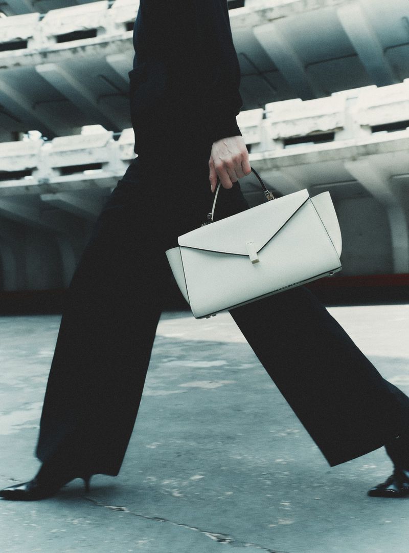 Model walking in dark trousers and sweater carrying a structured white handbag against an architectural background