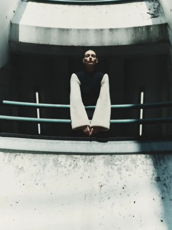 Model leaning on a teal railing against a weathered concrete architectural background with dramatic shadows.