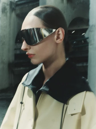 Model wearing futuristic silver visor sunglasses and a light yellow coat with a dark collar against a stone architectural background
