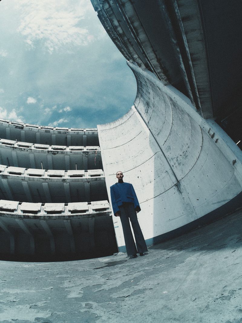 Model wearing a blue jacket and dark trousers standing inside a curved concrete architectural structure under a blue sky
