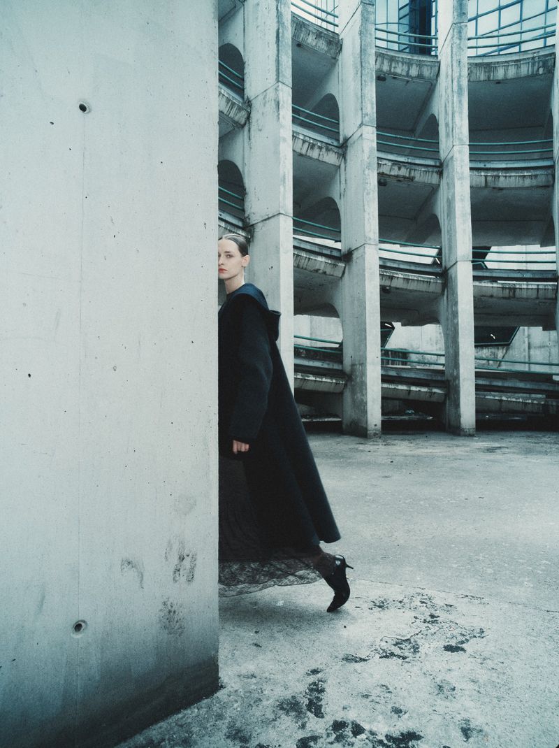 Model Daga Ziober wearing a long dark coat standing in a brutalist architectural structure with curved ramps.