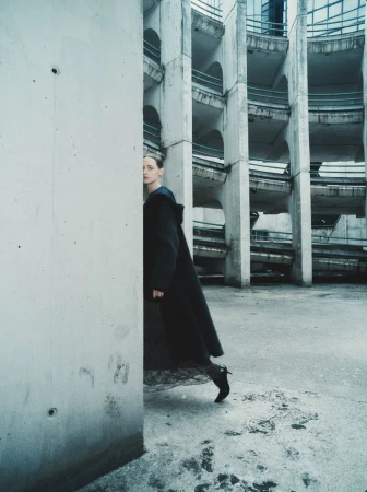 Model Daga Ziober wearing a long dark coat standing in a brutalist architectural structure with curved ramps.