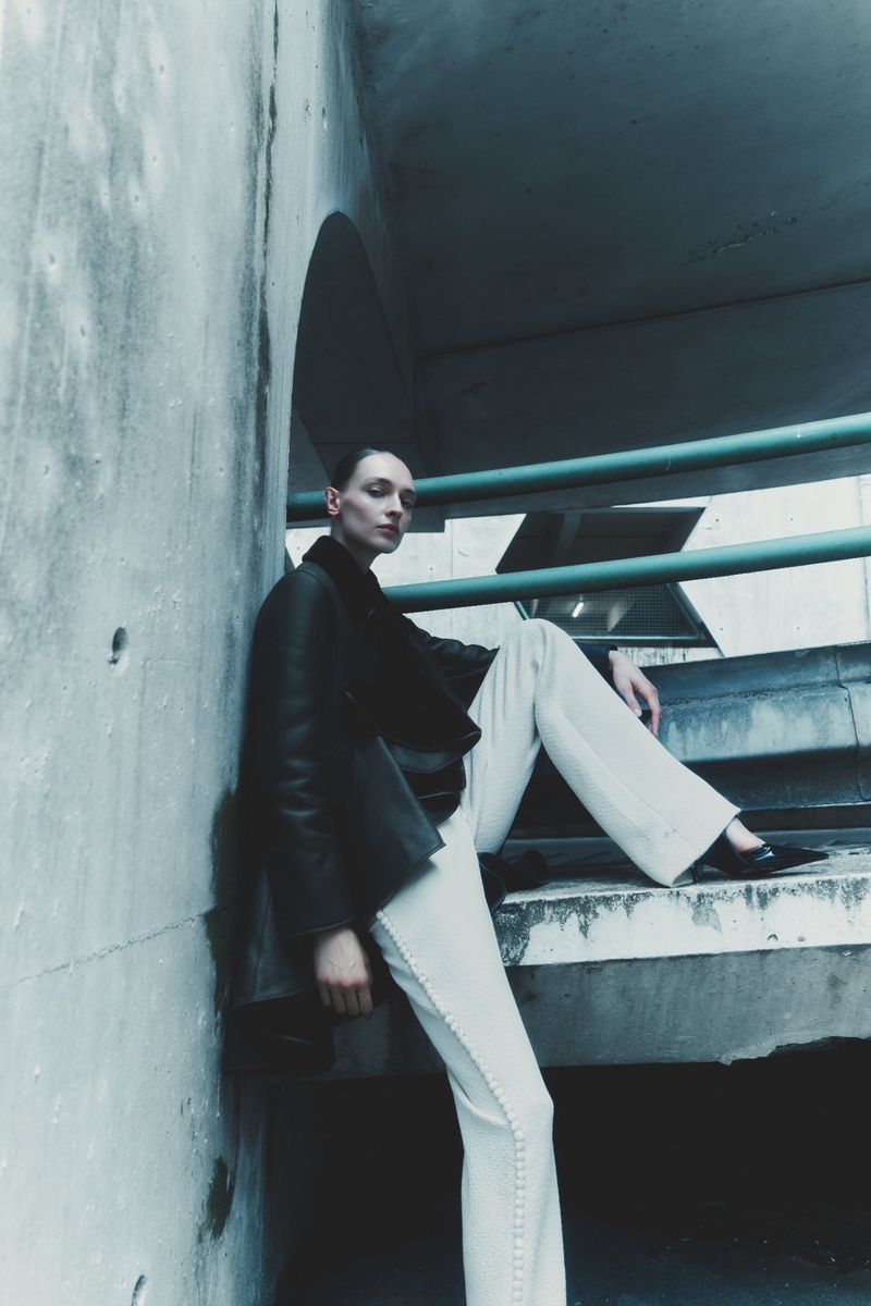 Daga Ziober wearing a dark jacket and white trousers seated in a concrete architectural structure for Seduction Magazine.