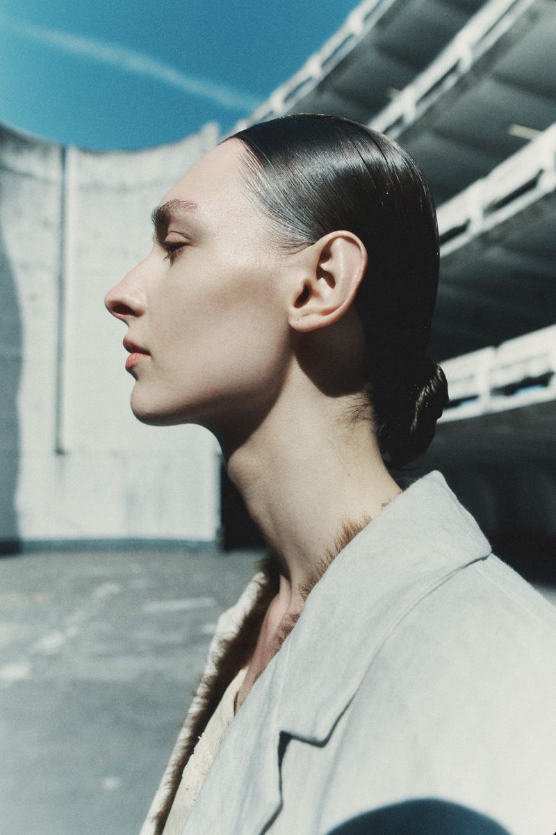 Profile portrait of a model with sleek dark hair in a bun wearing a light-coloured jacket against an architectural background