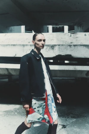 Model Daga Ziober wearing a dark jacket and printed skirt standing outdoors against a concrete architectural backdrop.