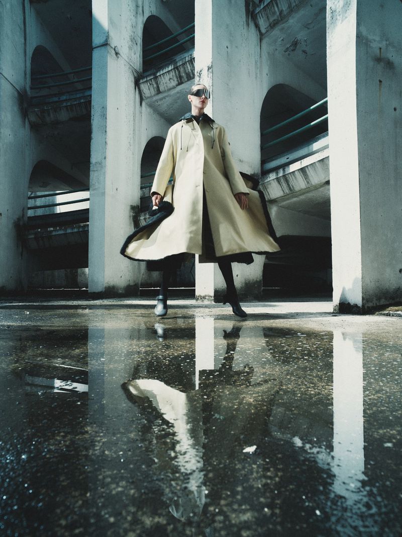Model wearing a long beige coat and sunglasses posing within a brutalist concrete structure with water puddle reflection