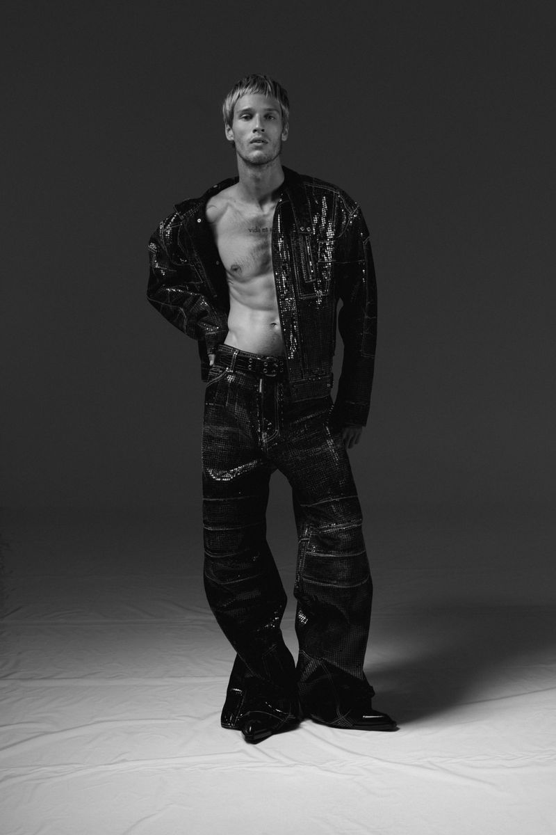 Monochrome fashion portrait of a male model wearing a dark glossy jacket and wide trousers on seamless white studio backdrop