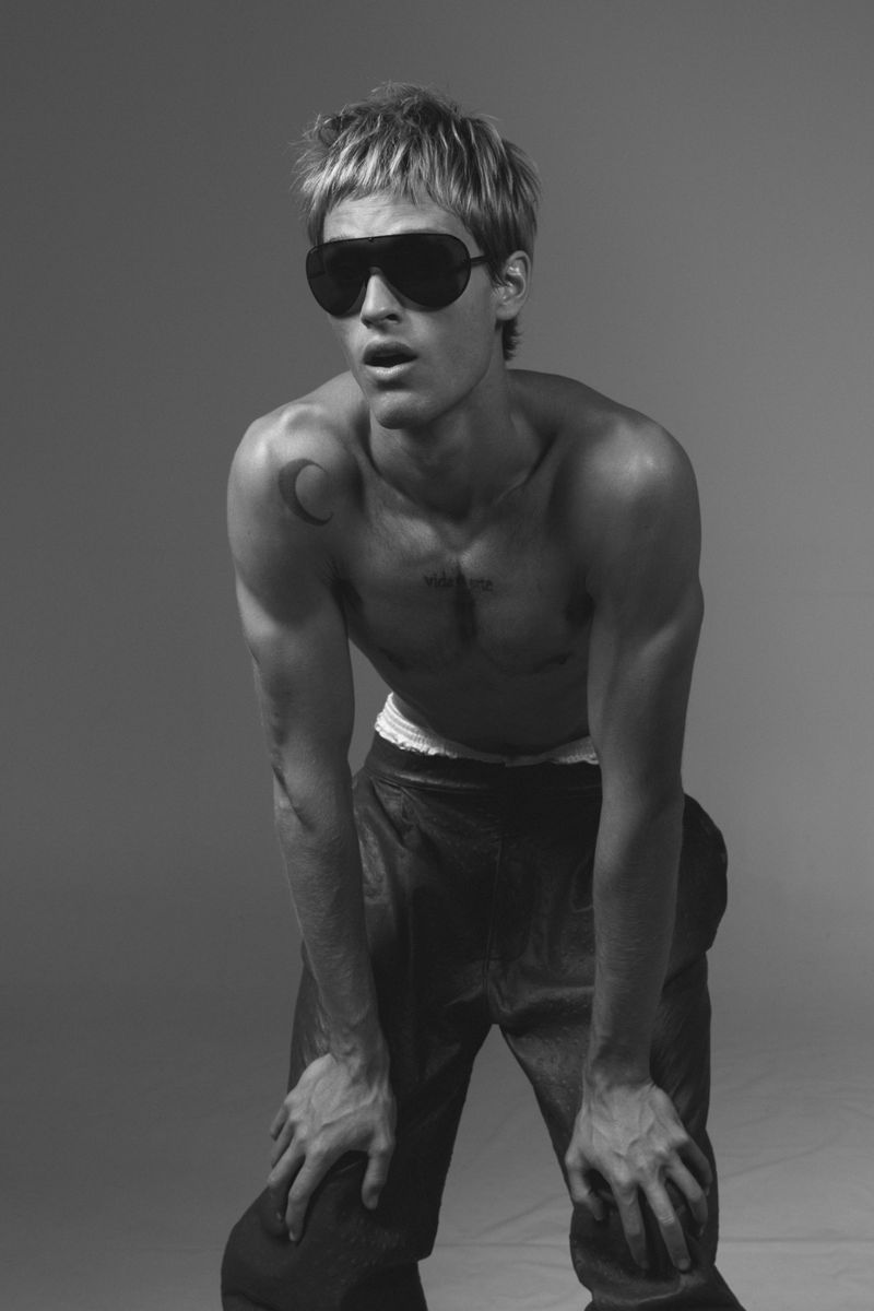 Monochrome portrait of male model Yeray Allgayer wearing dark sunglasses and dark trousers against a grey backdrop for Numero Netherlands.
