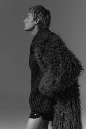 Profile portrait of a male model wearing a textured fluffy coat and ribbed sweater against a grey studio backdrop