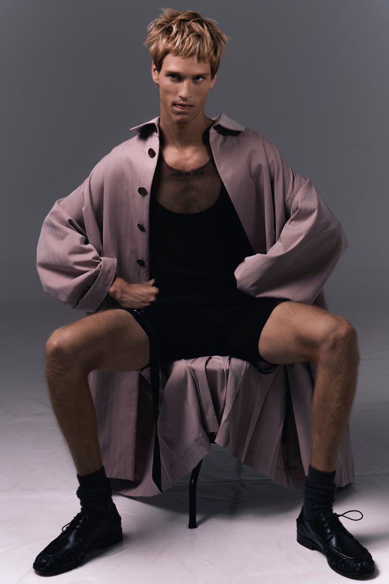 Model Yeray Allgayer seated in an oversized coat and black shorts against a grey studio backdrop