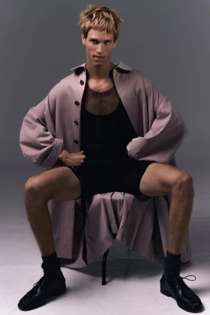 Model Yeray Allgayer seated in an oversized coat and black shorts against a grey studio backdrop