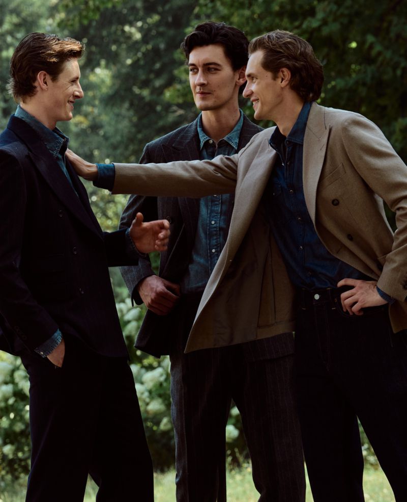 Three men in tailored blazers and denim shirts talking together outdoors in a green garden setting for Gentleman Magazine.