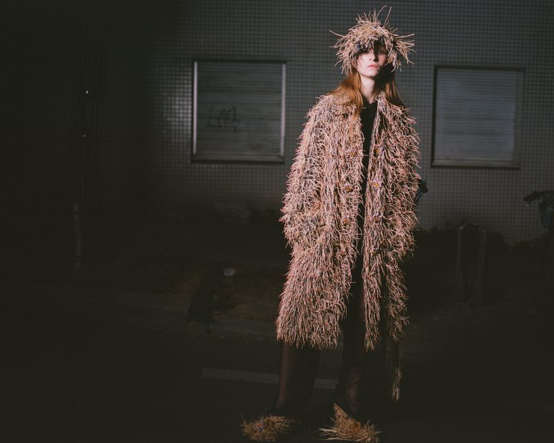 Model standing outdoors at night wearing a textured fringed straw coat and matching hat under dim lighting.