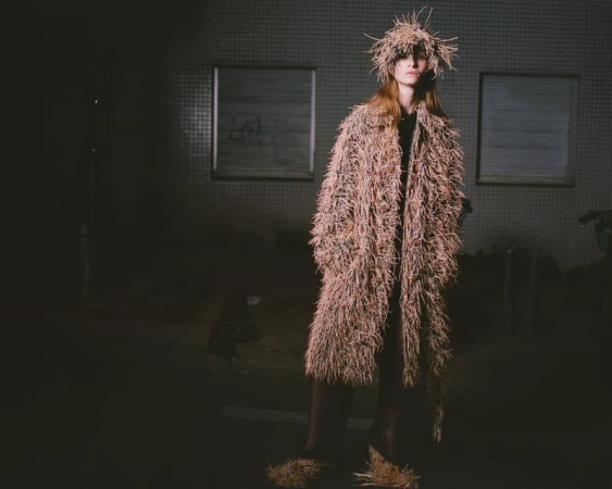 Model standing outdoors at night wearing a textured fringed straw coat and matching hat under dim lighting.