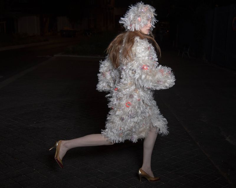 Model in a textured white fringed coat and matching hood running across a dark street at night wearing heels