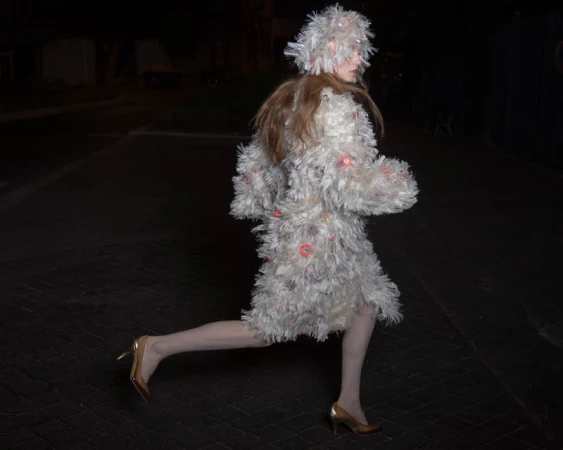 Model in a textured white fringed coat and matching hood running across a dark street at night wearing heels
